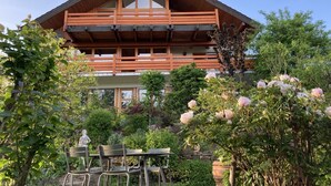 A two-story wooden house with a balcony, surrounded by lush greenery and flowering plants.
