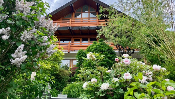 A multi-story building with a wooden facade, surrounded by lush greenery and flowering plants.