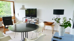 A modern dining area with a round table, white chairs, a television, books, and a plant.