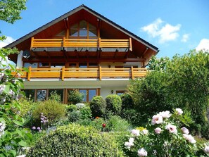 A two-story wooden chalet with balconies, surrounded by lush gardens and flowering plants.