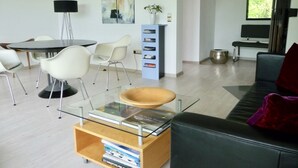 A modern living room with a glass coffee table, a black sofa, a dining table with chairs, a bookshelf, and a television.