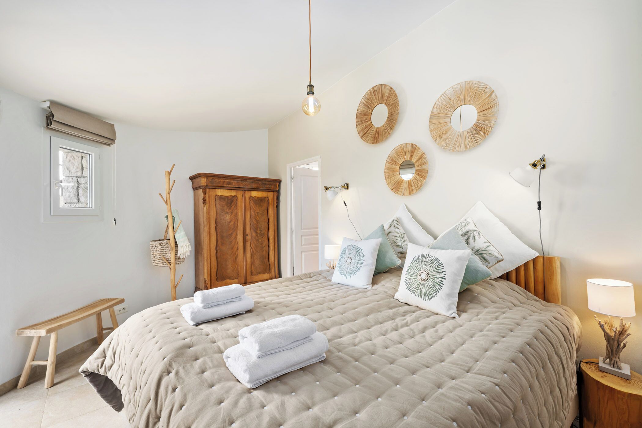 A bedroom with a large bed, a wooden wardrobe, a bench, and a bedside table with a lamp.