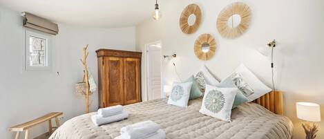 A bedroom with a large bed, a wooden wardrobe, a bench, and a bedside table with a lamp.