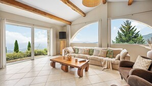 A spacious living room with a large wooden coffee table, a beige sofa with patterned cushions, and a view of the mountains through large windows.