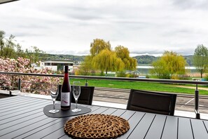 A table with a bottle of wine and a glass, overlooking a lake with trees and a distant building.