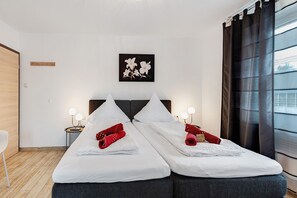 A double bed with white linens, two red towels, a wall-mounted artwork, bedside lamps, and a window with dark curtains.