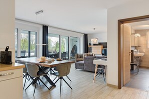 A modern kitchen with a dining area, large windows, and a view of greenery outside.