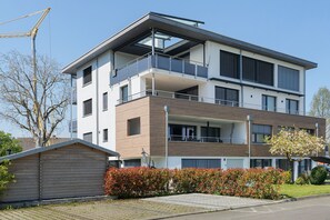 A modern multi-story residential building with a balcony, a garage, and a well-maintained garden.