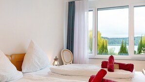 Habitación de hotel con dos camas, ropa de cama blanca, una mesita de noche con lámpara, ventana con vistas a árboles y agua, y toallas rojas sobre las camas.