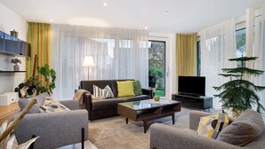 En moderne stue med sofa, lænestole, sofabord, fjernsyn og et stort vindue med gardiner.
