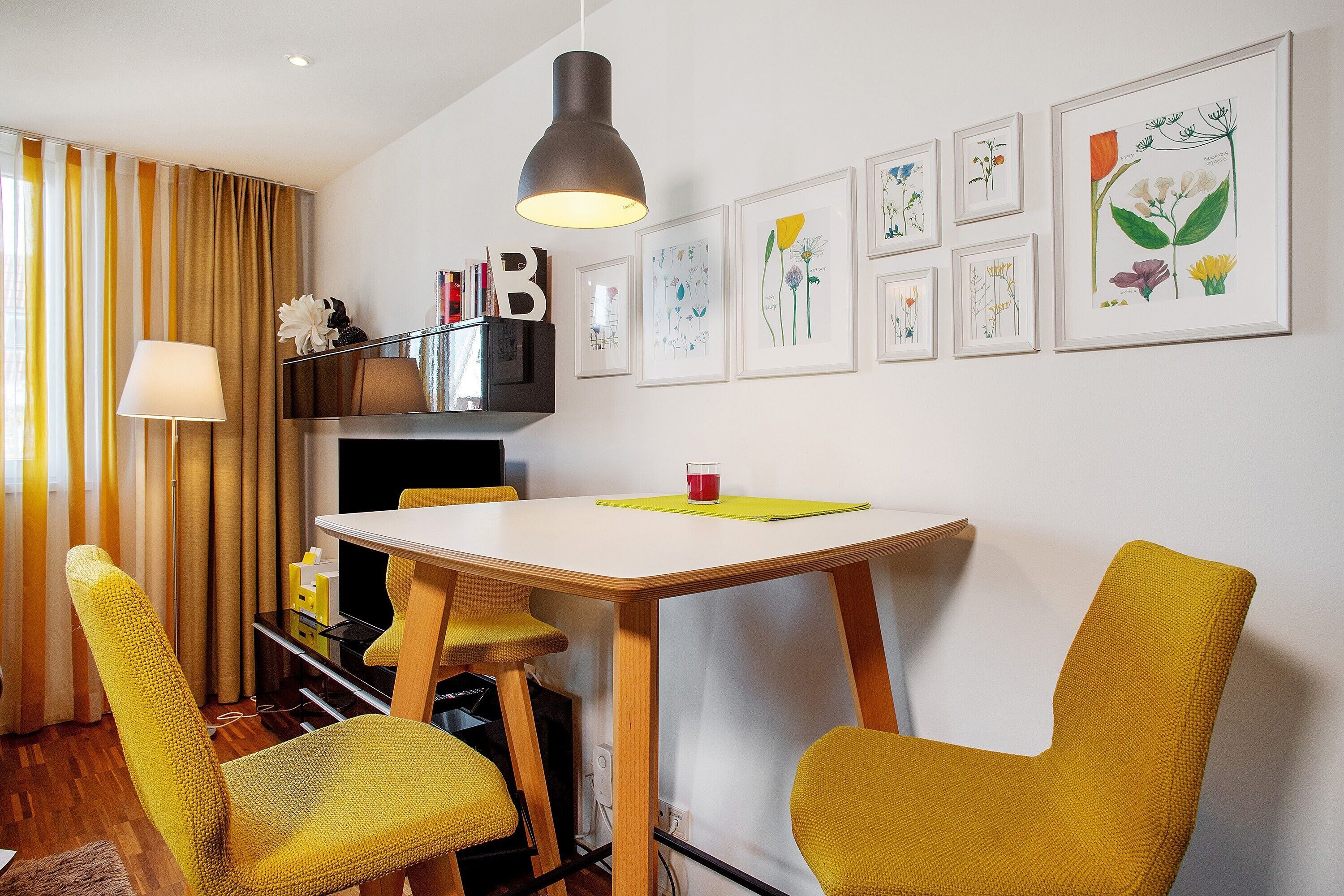 Ein modernes Zimmer mit Esstisch, gelben Stühlen, einer Pendelleuchte und gerahmten Pflanzenmotiven an der Wand.