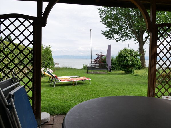 A patio with a table and chairs, a lawn chair, and a view of a body of water. - Haus Alpenblick – Happiness Apartment with Balcony and Wi-Fi (Immenstaad am Bodensee)