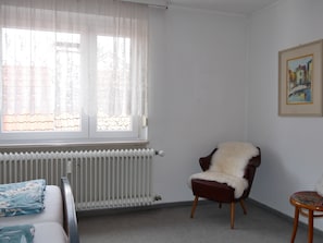 A room with a radiator, a chair with a fur throw, a small stool, and a framed painting on the wall. - Haus Alpenblick – Happiness Apartment with Balcony and Wi-Fi (Immenstaad am Bodensee)