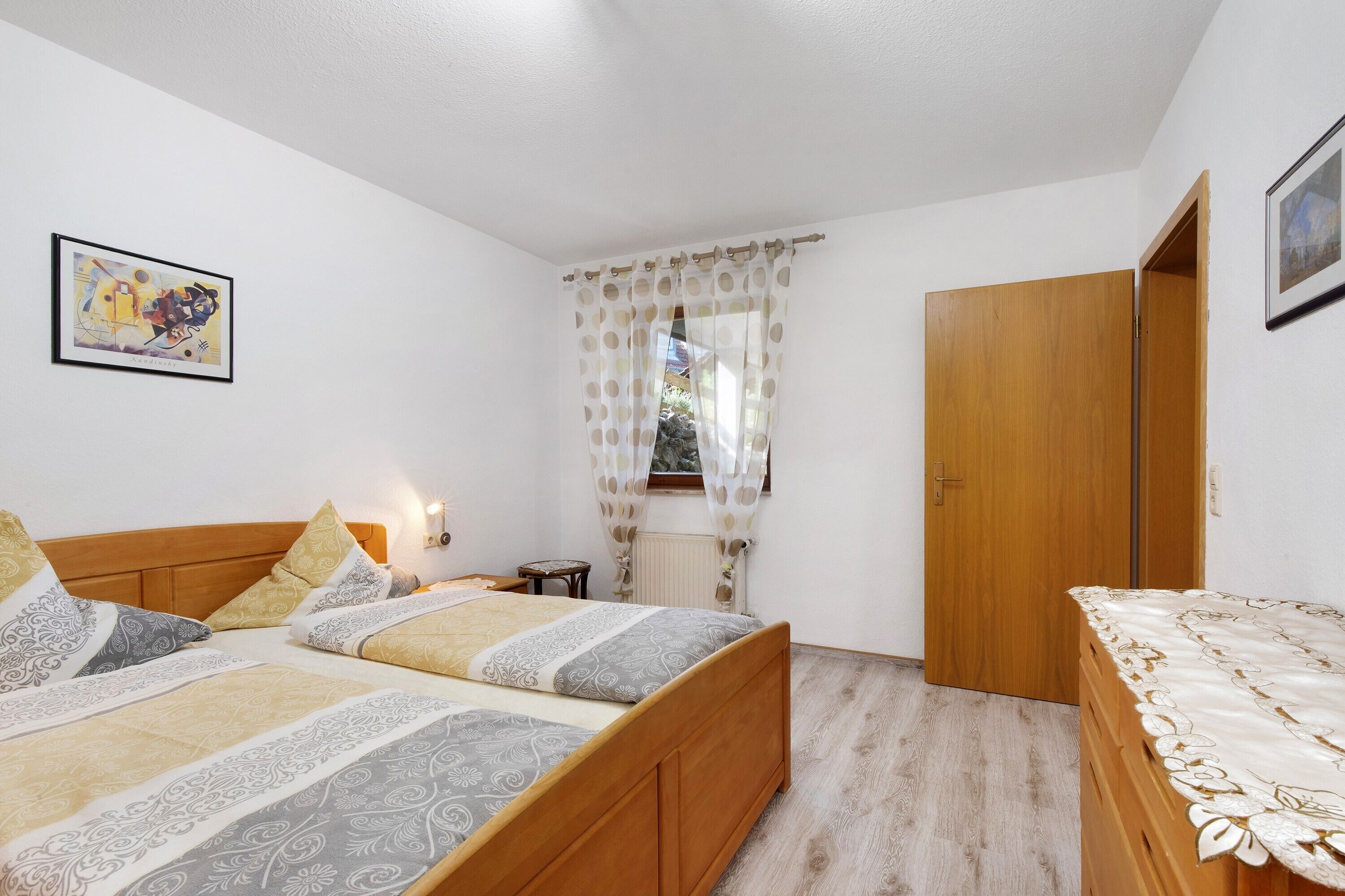 A bedroom with a bed, a bedside table, a lamp, a window with curtains, and a door.