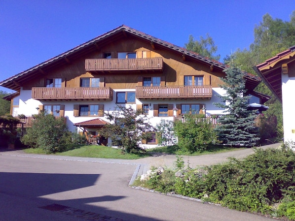 A multi-story wooden building with balconies, a paved driveway, and surrounding greenery.