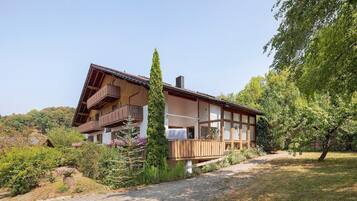 A two-story chalet with a sloped roof, wooden balconies, and a garden with various plants and flowers.