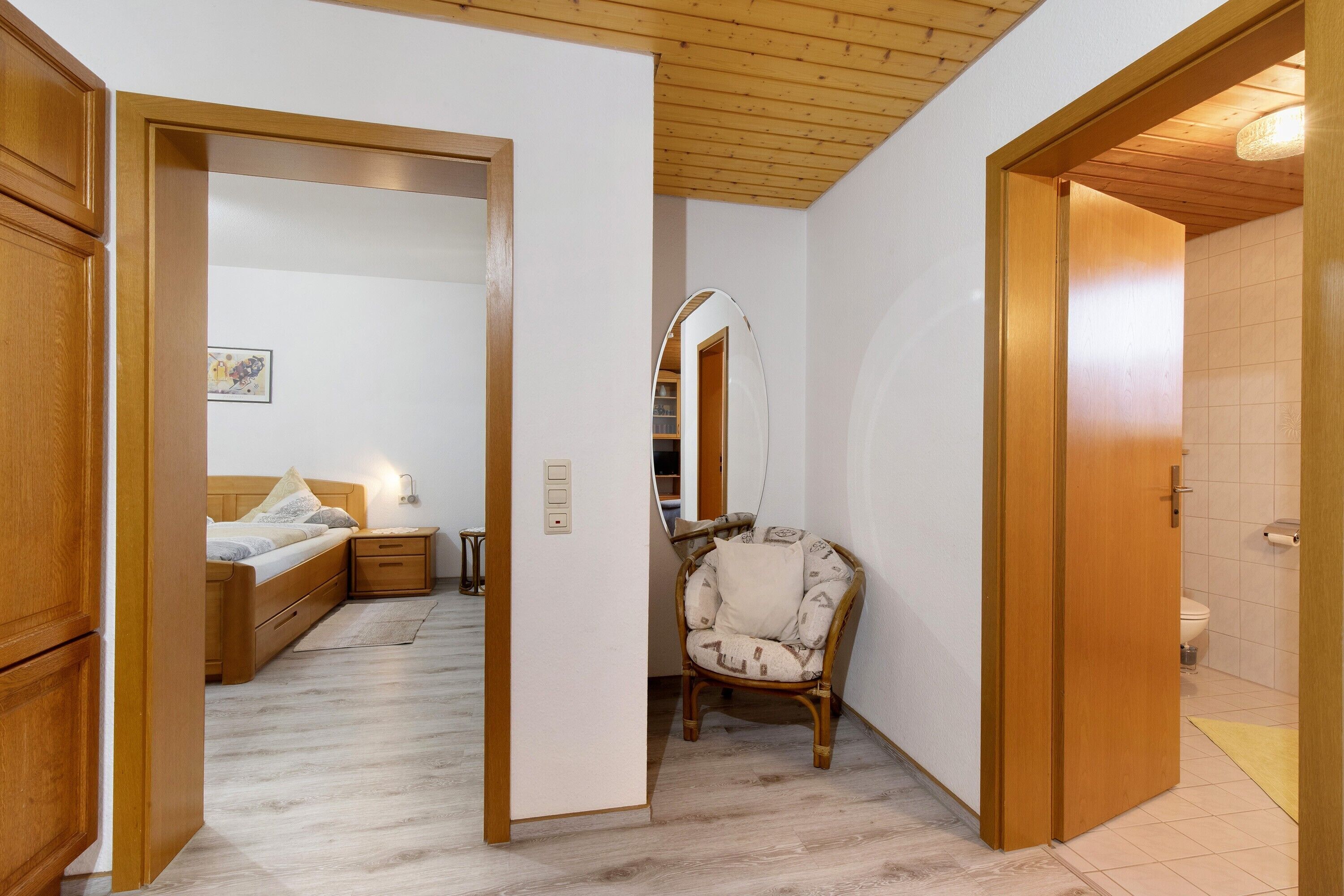 Ein Schlafzimmer mit Bett, Nachttisch und Stuhl. Es gibt ein Badezimmer mit Toilette und Spiegel.