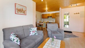 Ein Wohnzimmer mit grauem Sofa, einem passenden Sessel und einem Couchtisch mit einer Vase. Im Hintergrund ist eine Essecke mit Tisch und Stühlen zu sehen.