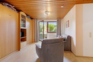 A room with wooden flooring, a grey sofa, a television on a cabinet, and a window with a view of greenery. - Ferienwohnung 'B' mit privater Terrasse und WLAN (Lonsee)
