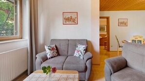 Ein Wohnzimmer mit einem grauen Sofa, zwei blumigen Kissen, einer Tischläufer aus Spitze und einer Vase mit einer einzelnen Blume. An der Wand befinden sich zwei gerahmte Kunstwerke.