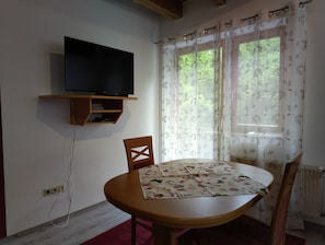 Een kamer met een televisie op een plank, een ronde houten tafel, stoelen en een raam met bloemen gordijnen.