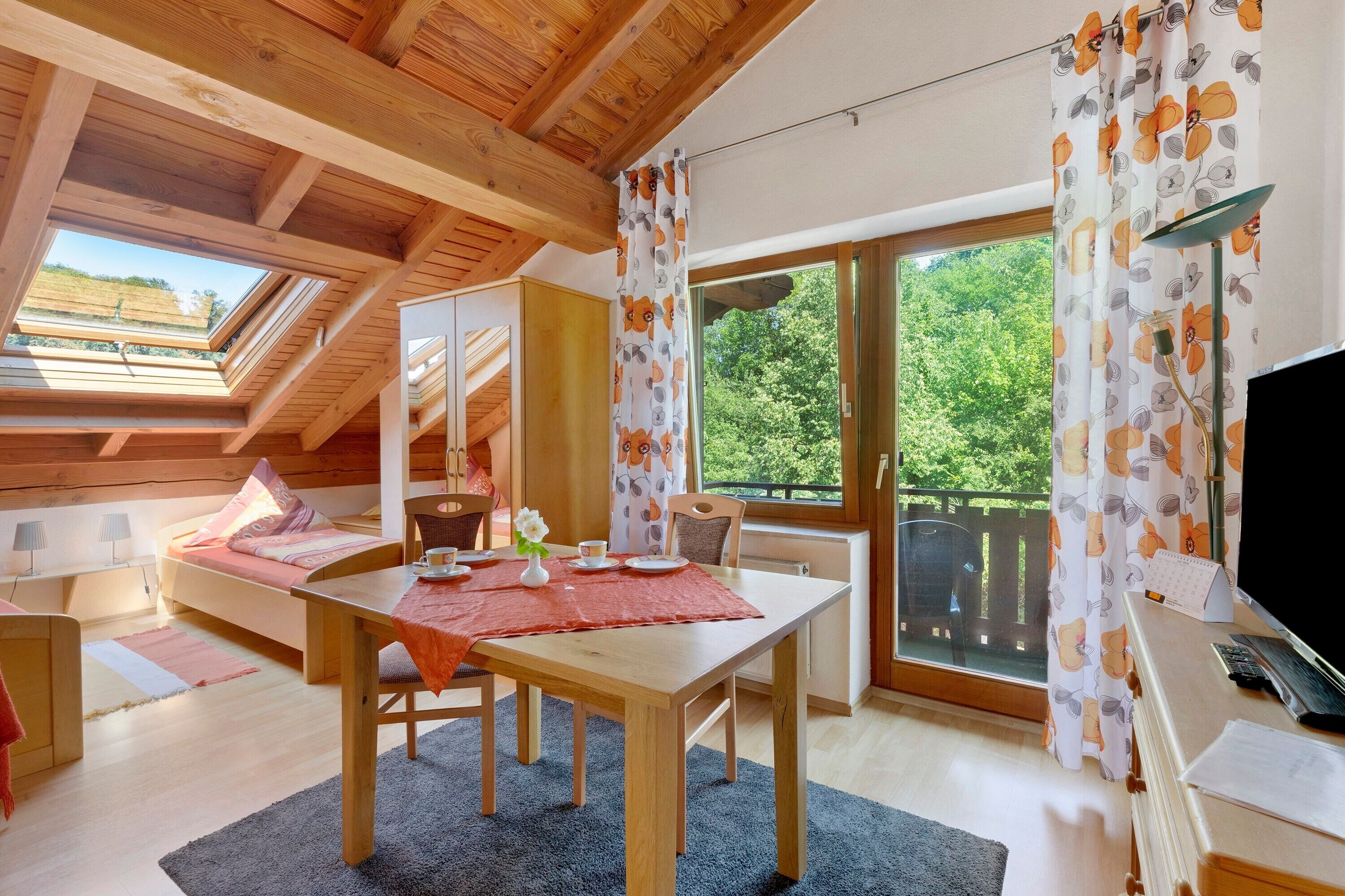 A cozy room with a wooden ceiling, a dining table with chairs, a TV, and large windows with floral curtains.