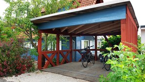 Una terraza cubierta de madera con una bicicleta, rodeada de follaje y un edificio de fachada azul.
