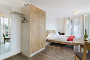 Een moderne hotelkamer met een houten garderobe, een bed met witte lakens, een eettafel met een fles en glas, en uitzicht door het raam.