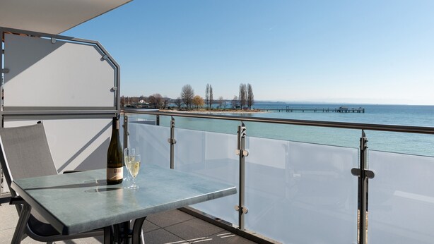 En balkon med et glasbord, en flaske hvidvin og frit udsyn til havet og en mole i baggrunden.