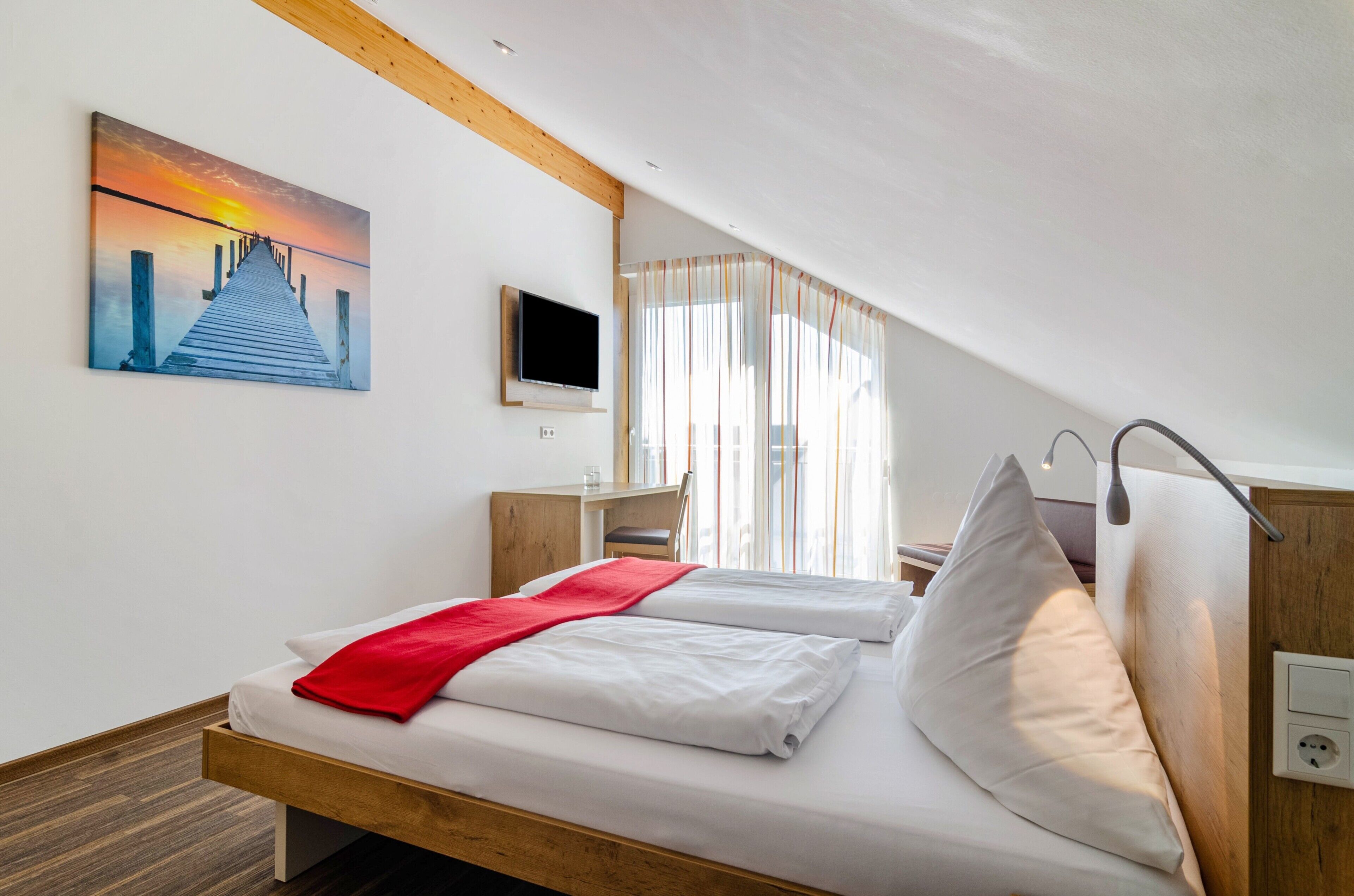 A hotel room with two beds, a TV, a desk, and a painting of a sunset over a pier.