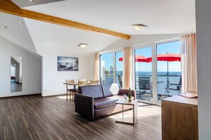 Ein modernes Wohnzimmer mit einer Couch, einem Couchtisch und einem Essbereich, große Fenster mit Meerblick und ein Balkon mit roten Sonnenschirmen.