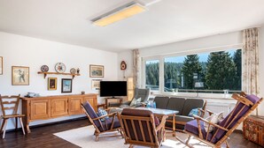 Een woonkamer met een houten kast, een televisie, een bank en een groot raam met uitzicht op bomen.