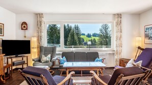 Een woonkamer met een groot raam, een televisie en zitplaatsen.