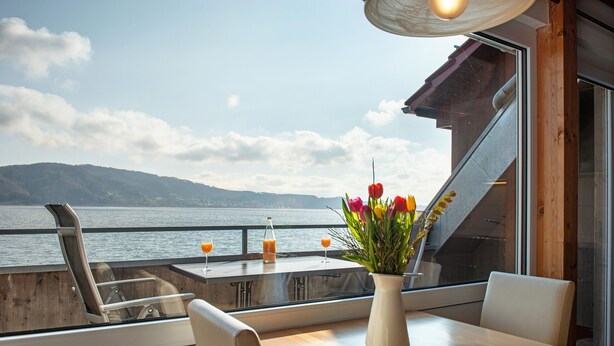 A dining area with a view of the water, a table with chairs, a vase with flowers, and a bottle of orange juice. - Apartment '8, 53 sqm, Directly on Lake Constance with Lake Access (Bodman-Ludwigshafen)