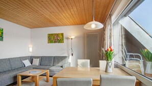 A modern living room with a wooden ceiling, a grey sofa, a wooden dining table, and a large window with curtains. - Apartment '8, 53 sqm, Directly on Lake Constance with Lake Access (Bodman-Ludwigshafen)