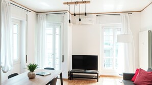 A modern living room with a wooden dining table, a flat-screen TV on a stand, a grey sofa with red cushions, and large windows with white curtains. - Suite Duomo View 47 (Como)