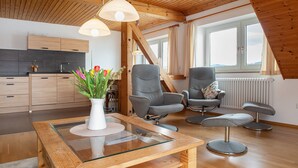A modern loft with a wooden ceiling, a kitchen with dark cabinets, two grey chairs, a glass table, and a view of the sky through a large window. - Apartment '7, 62sqm' with Large Sunny Balcony and Direct Lake Access (Bodman-Ludwigshafen)
