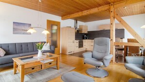 A modern living room with a sofa, a coffee table, and a grey armchair. There is a kitchen area with wooden cabinets and a dining table. - Apartment '7, 62sqm' with Large Sunny Balcony and Direct Lake Access (Bodman-Ludwigshafen)