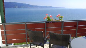 A balcony with wicker chairs, a table, and a view of a calm sea and distant hills. - Apartment '7, 62sqm' with Large Sunny Balcony and Direct Lake Access (Bodman-Ludwigshafen)