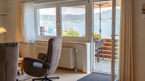 A room with a large window, a chair, a table, and a view of a body of water and hills.
