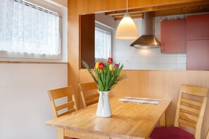 Una mesa de comedor de madera con un jarrón blanco de flores, sillas de madera, y una cocina con armarios y una lámpara colgante.