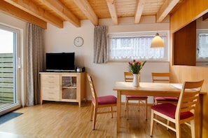 Une pièce avec un plafond en bois, une table à manger avec des chaises, un téléviseur posé sur un meuble et une fenêtre avec des rideaux.