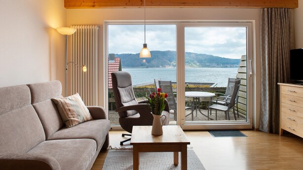 Ein modernes Wohnzimmer mit einer Couch, einem Sessel, einem Tisch mit einer Vase und Blick auf das Meer durch große Glasfenster.