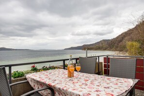 A table with a floral tablecloth, two glasses of orange juice, and a bottle of juice on a balcony overlooking a body of water.