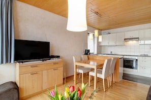 A modern kitchen with a flat-screen TV, wooden cabinets, a dining table with chairs, and a wooden ceiling.