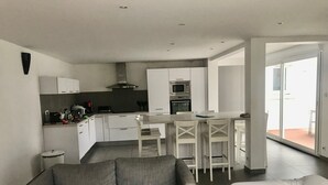 Eine moderne Küche mit weißen Schränken, ein Essbereich mit Tisch und Stühlen, ein graues Sofa und ein Bett mit weißer Bettwäsche.