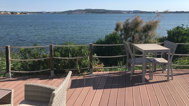 Eine Holzterrasse mit Tisch und Stuhl mit Blick auf ein Gewässer, in der Ferne Land und ein Leuchtturm.