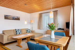Un salón moderno con techo de madera, un sofá, una mesa de centro, una mesa de comedor con sillas, un televisor y una ventana con cortinas.