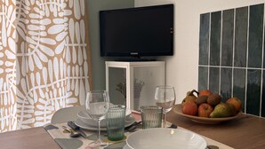 Un comedor con una mesa de madera dispuesta para dos personas, un reloj en la pared, un televisor montado sobre la mesa y un frutero en la mesa.
