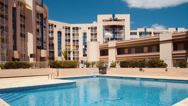 Una piscina con superficie de agua azul cristalina, rodeada por un deck empedrado. En el fondo, se observa un edificio de varios pisos con un diseño arquitectónico moderno, con balcones y grandes ventanales.